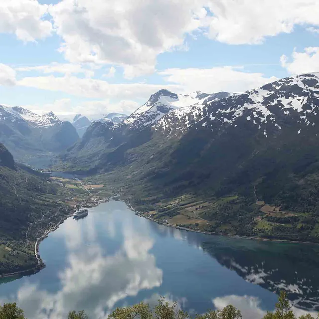Utsikt fra fjelltopp ned over en dyp dal med en blank fjord som speiler himmelen.