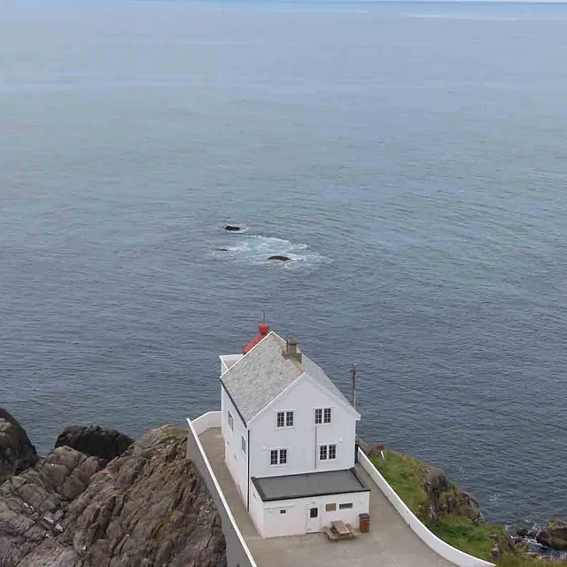 Et hvitmalt fyr eller hus på en bratt klippe mot åpent hav.