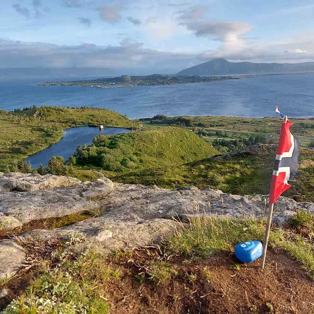 Utsikt fra en høyde med et norsk flagg, over et grønt kystlandskap med øyer og hav.
