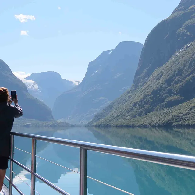 En person står på dekk av en båt og fotograferer en dyp fjord omkranset av høye fjell.