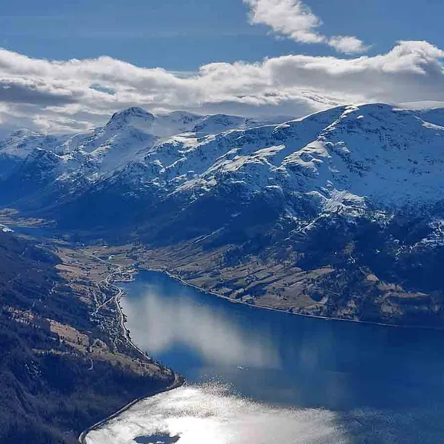Panoramautsikt fra fjellet over en dyp, blå fjord omkranset av snødekte fjelltopper under en delvis skyet himmel.
