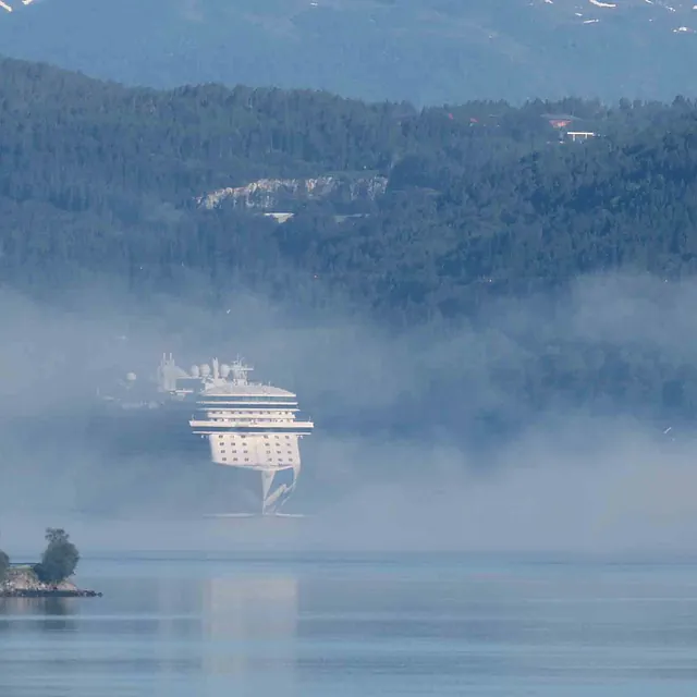 Et stort cruiseskip delvis skjult av tett lavtliggende tåke på fjorden.