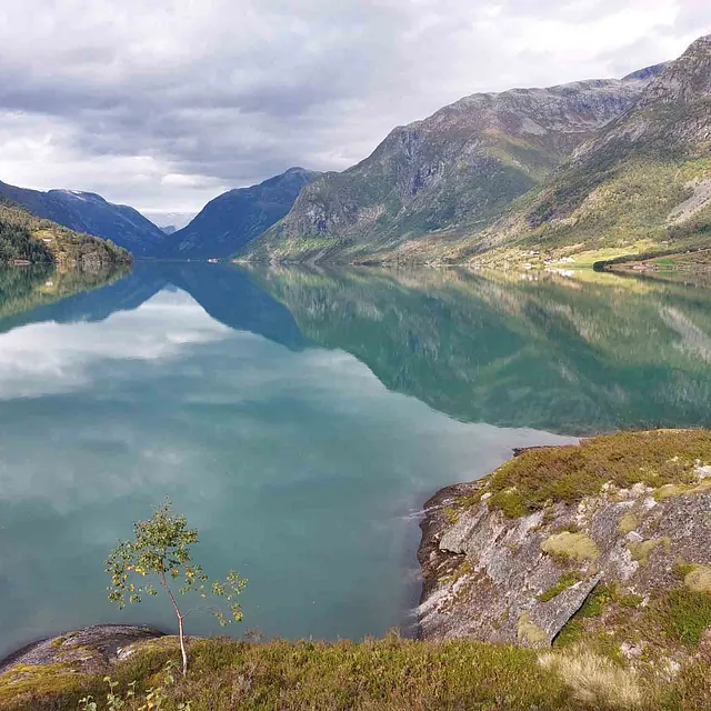 Blikkstille, grønn innsjø som reflekterer bratte fjellsider og en skyet himmel.