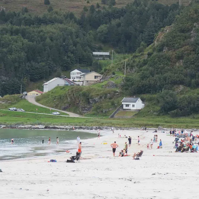 Sandstrand med mennesker, hvitmalt hus og grønne åser.