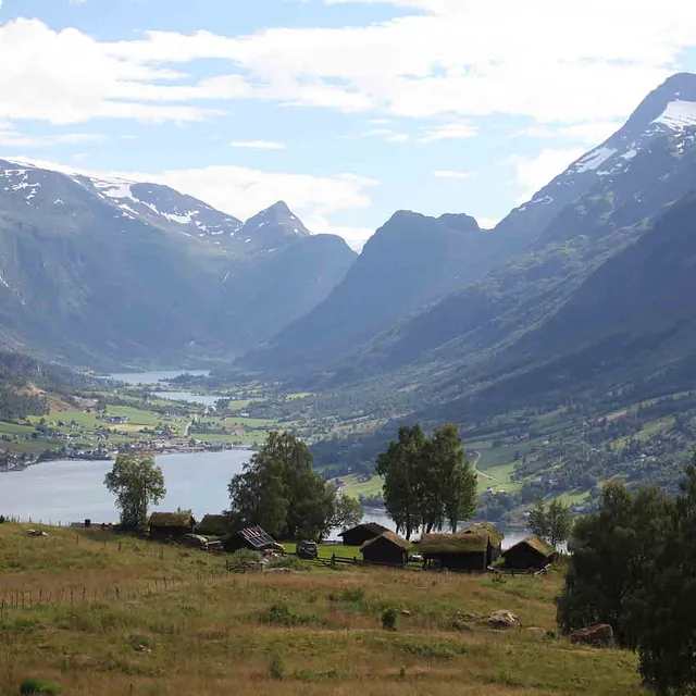 Utsikt fra en gressbakke ned mot en dyp dal med et vann og høye, bratte fjell i bakgrunnen.