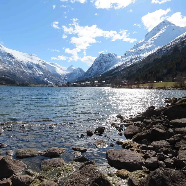 Steinete strandkant ved et klart vann i en dyp fjelldal.