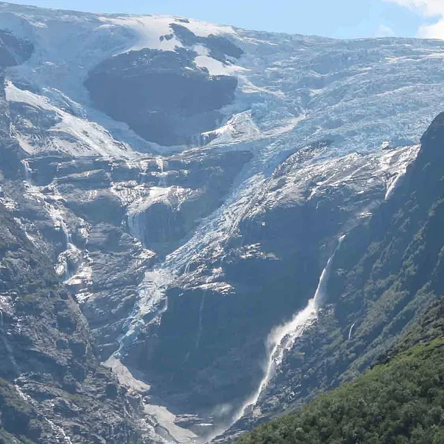 Snø- og isdekt fjellside som reiser seg over en grønnkledd dalbunn.