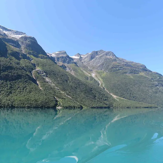 Klart, speilblankt turkis brevann som reflekterer høye, grønne fjell under en blå himmel.